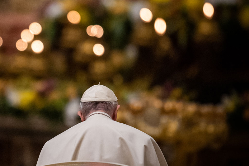 Pope Francis, with his back to the camera, faces a wall of candles. He wears white vestments and bows his head.